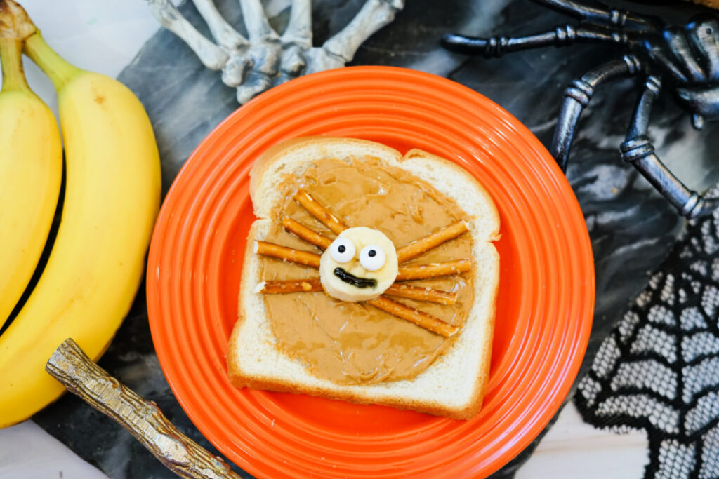 Halloween Toast That'll Make You Go BOO-nanas 6 A slice of Halloween Toast with peanut butter, topped with banana slices, pretzel stick legs, and candy eyes to resemble a spider, served on an orange plate surrounded by bananas and festive Halloween decorations.