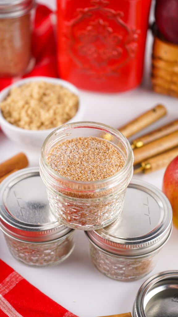 A small glass jar filled with spiced brown sugar, perfect for an Apple Cider Mix, sits atop two closed jars, surrounded by cinnamon sticks and other jars on a white surface.
