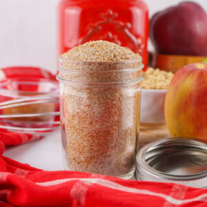 A glass jar filled with brown granulated sugar sits on a white surface, surrounded by a red cloth, apples, an Apple Cider Mix container, and kitchen utensils in the background.