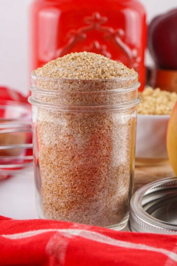 A glass jar filled with brown granulated sugar sits on a white surface, surrounded by a red cloth, apples, an Apple Cider Mix container, and kitchen utensils in the background.