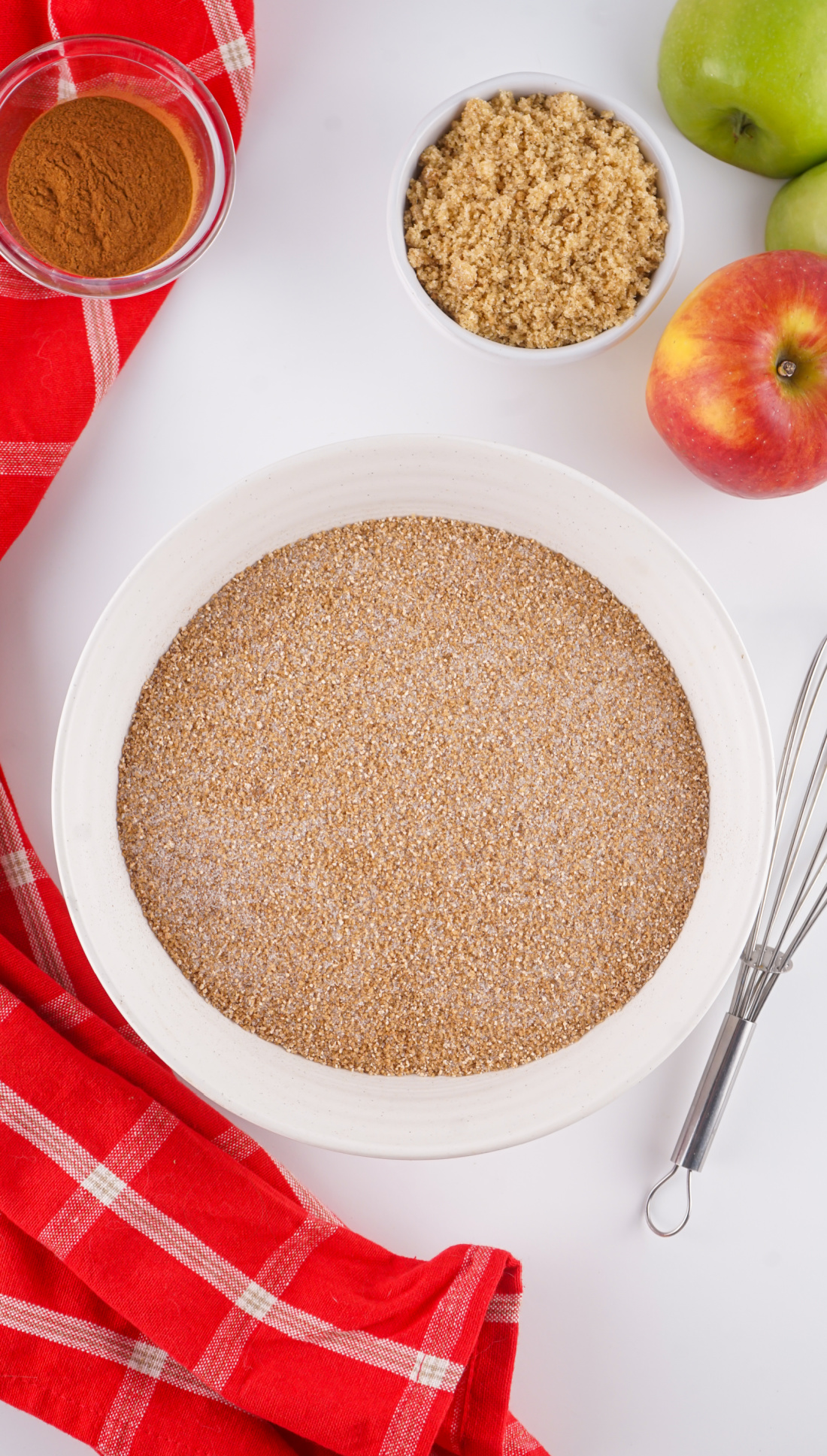 A white bowl filled with a brown sugar and spice mixture-perfect for your Apple Cider Mix-sits on a white surface beside apples, a bowl of brown sugar, cinnamon, a whisk, and a red checkered cloth.