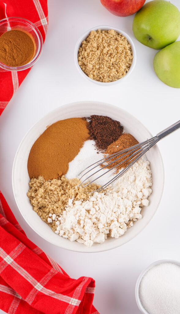 A bowl with flour, brown sugar, white sugar, cinnamon, nutmeg, and other dry ingredients sits next to whole green apples and bowls of apple cider mix and cinnamon, with a whisk and red cloth nearby.