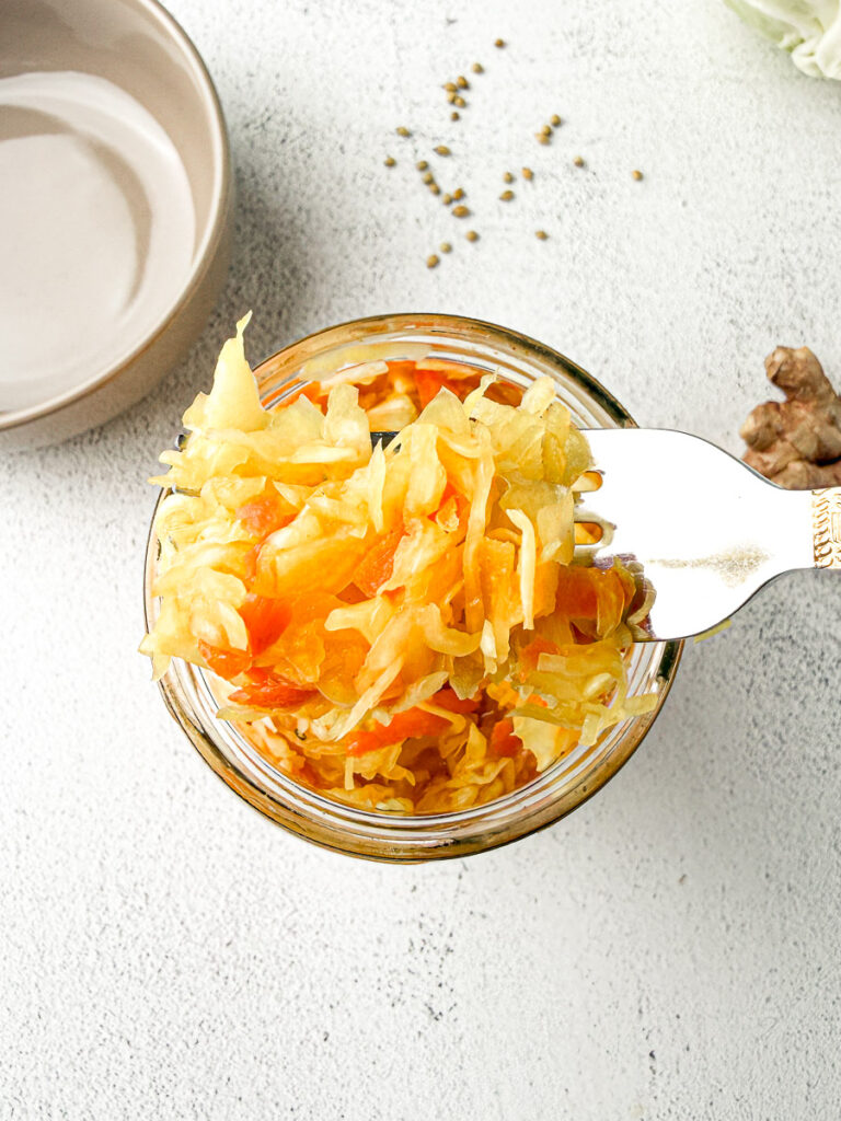 Easy Small-Batch Sauerkraut with Turmeric & Mustard 2 Overhead view of vibrant golden small-batch sauerkraut, colored with turmeric, in a mason jar on a light countertop with a forkful of it next to empty brown bowl, ginger and cabbage leaves.