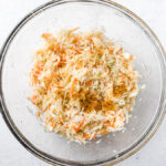 Fresh shredded cabbage and carrots with turmeric, mustard powder, black pepper and salt for small-batch golden sauerkraut in a large glass bowl on light countertop.