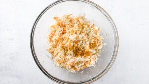 Fresh shredded cabbage and carrots with turmeric, mustard powder, black pepper and salt for small-batch golden sauerkraut in a large glass bowl on light countertop.