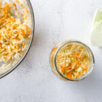 Fresh shredded cabbage for small-batch golden sauerkraut in a large glass bowl on light countertop next to cabbage leaves and mason jar with sauerkraut.