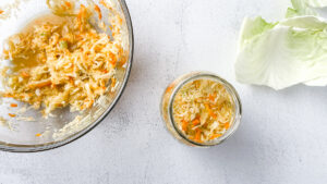 Fresh shredded cabbage for small-batch golden sauerkraut in a large glass bowl on light countertop next to cabbage leaves and mason jar with sauerkraut.