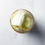Fresh shredded cabbage and carrots with turmeric, mustard powder, black pepper and salt for small-batch golden sauerkraut in a glass mason jar covered with cabbage leaves on light countertop.