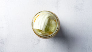 Fresh shredded cabbage and carrots with turmeric, mustard powder, black pepper and salt for small-batch golden sauerkraut in a glass mason jar covered with cabbage leaves on light countertop.