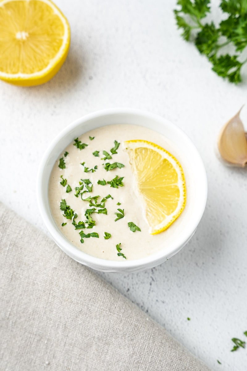 creamy lemon tahini sauces in white bowl with a lemon slice and parsley on top