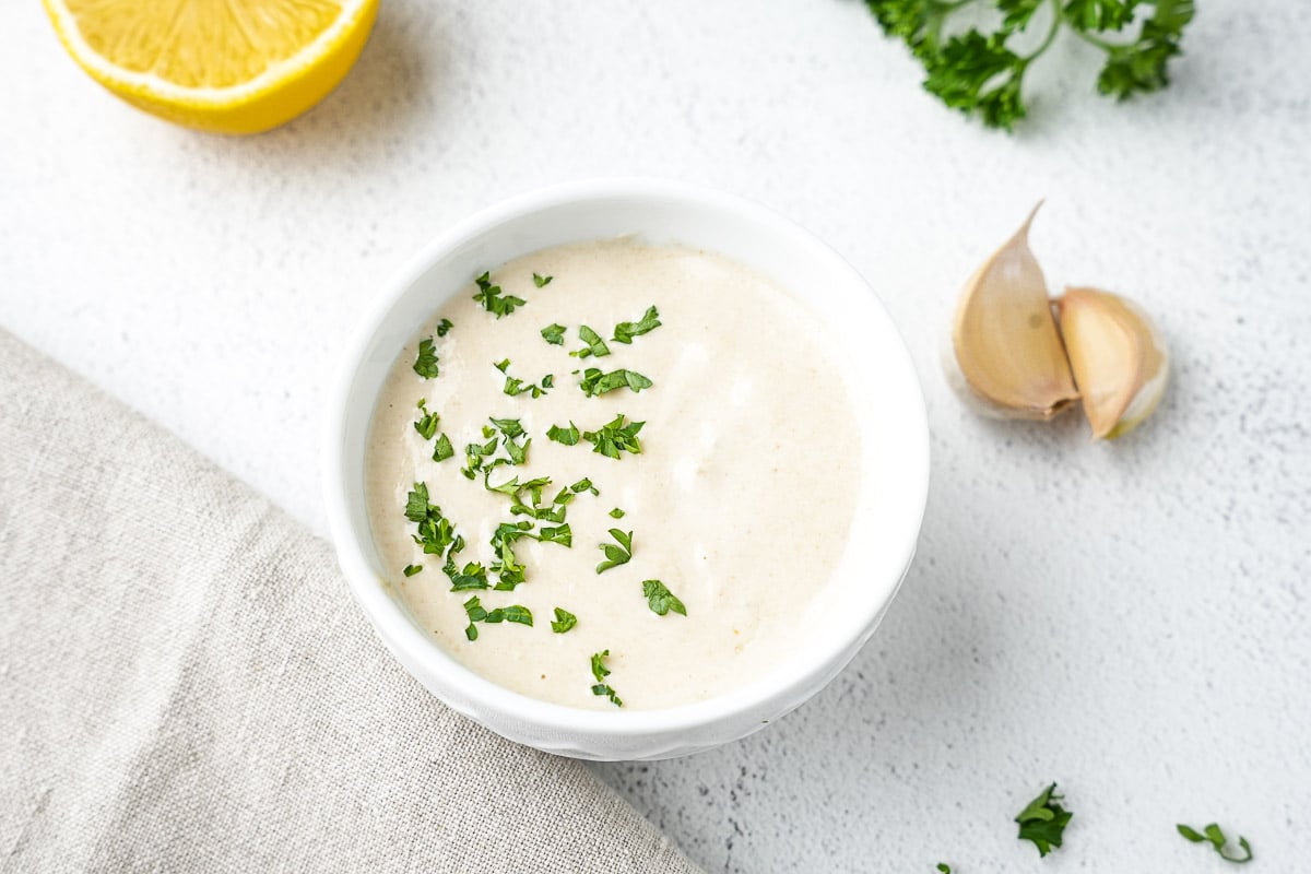 Creamy lemon tahini sauce in bowl with garlic cloves next to it, lemon and parsley