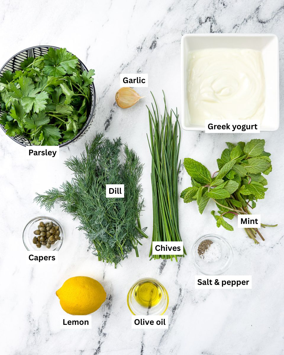 Green goddess dressing recipe ingredients on countertop: parsley, garlic clove, greek yogurt, mint, chives, dill, capers, lemon, olive oil, salt & pepper