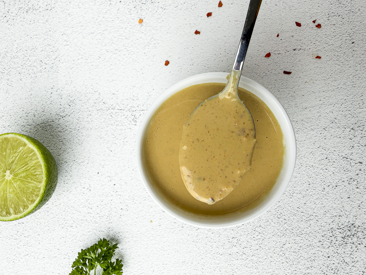 Spicy peanut satay sauce in a sauce bowl dripping from spoon next to a lime and parsley.